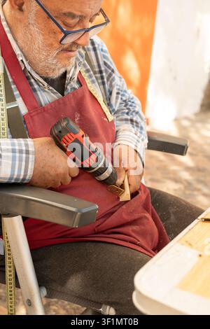 Close-up of an elderly craftsman wearing glasses and apron, using a cordless drill to fasten a metal bracket to wood, working carefully in Palamos. Stock Photo