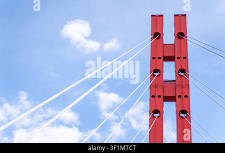 Low-angle view of a red suspension bridge tower with white cables stretching diagonally across a bright blue sky with soft clouds, highlighting modern Stock Photo