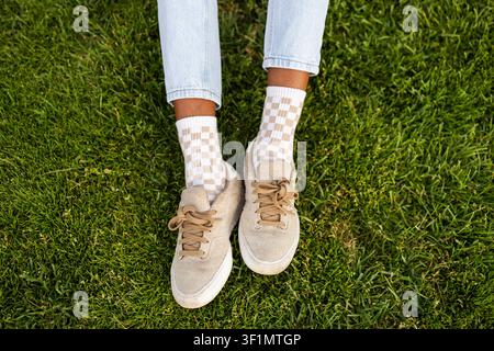 Top view of a person standing on fresh green grass wearing beige sneakers, checkered socks, and light jeans, creating a relaxed outdoor lifestyle fash Stock Photo