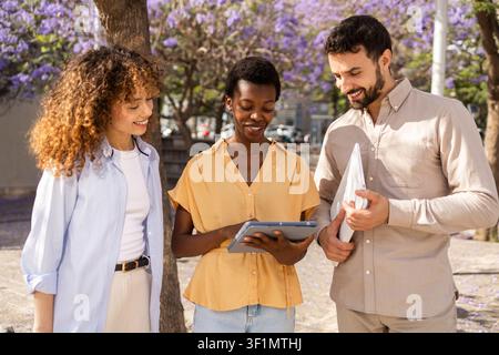 Three diverse colleagues collaborate outdoors, smiling while reviewing data on a tablet , with blooming purple trees creating a relaxed workplace mood Stock Photo