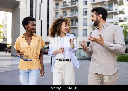 Three colleagues walk and talk in a modern residential courtyard, smiling and holding a laptop, discussing work and real estate in an urban apartment Stock Photo