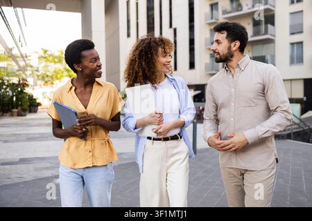 Three diverse coworkers walk outdoors in a modern urban district, chatting and smiling while carrying folders and a laptop, illustrating teamwork, bus Stock Photo