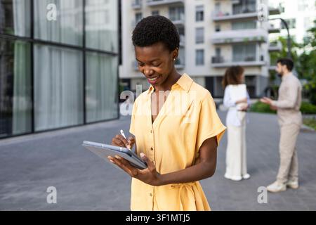 Smiling young woman in a yellow dress writes on a digital tablet outdoors in a modern urban courtyard, with office buildings and colleagues chatting i Stock Photo