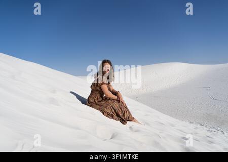 Beautiful woman traveler sitting on bright white sand dunes in the USA, enjoying a sunny day under a deep blue sky, with strong contrast between light Stock Photo