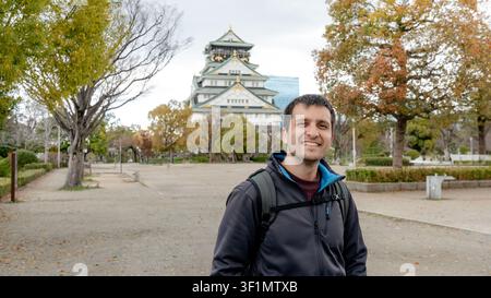 Smiling traveler standing in front of the majestic main tower of Osaka Castle, Japan, surrounded by serene parkland, showcasing stunning autumn foliag Stock Photo