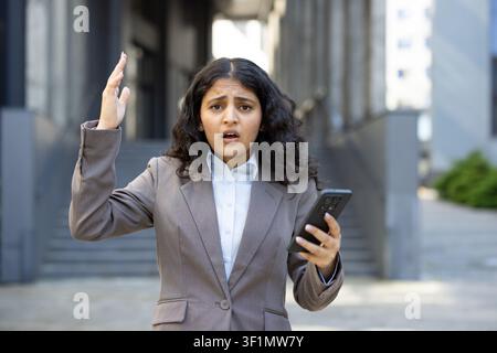 Businesswoman looking shocked and upset, raising her hand while reading disturbing information on her mobile phone outside a modern office building, expressing disbelief and stress Stock Photo