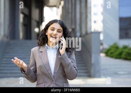 Happy young businesswoman having a lively conversation on a mobile phone outside an urban office building, gesturing with her hand while discussing business or personal topics outdoors Stock Photo