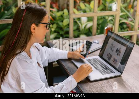 Woman working remotely with laptop and smartphone outdoors in tropical garden setting Stock Photo