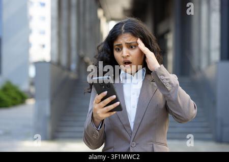 Young businesswoman with a shocked expression, holding a smartphone and looking at its screen with wide eyes, experiencing a moment of failure or a significant problem while standing outdoors Stock Photo