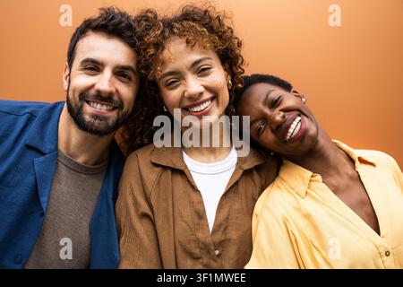 Close-up portrait of three diverse friends smiling and leaning together against a warm orange background, capturing friendship, inclusion, and joyful Stock Photo