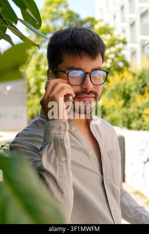 Young man with dark hair and eyeglasses talks on a smartphone outdoors near modern buildings and green plants, capturing casual communication, lifesty Stock Photo