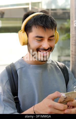 Smiling young man wearing yellow headphones and backpack checks a smartphone outdoors in an urban setting, enjoying music and messaging while walking Stock Photo