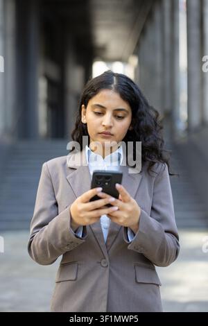 Young professional business woman standing outdoors, dressed in a smart suit, intently using her mobile phone for communication, browsing, or managing tasks Stock Photo