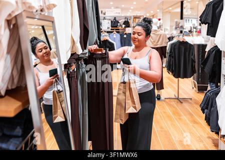 Asian woman browsing clothing racks in a modern fashion store, holding shopping bags and checking her smartphone, with mirror reflection and warm reta Stock Photo