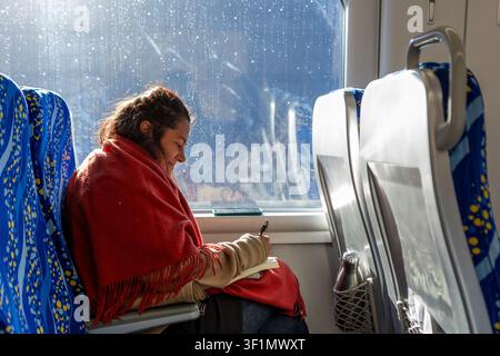 A woman wrapped in a red blanket sits by a train window, writing in a notebook. Sunlight and raindrops on the glass create a calm travel mood. Stock Photo