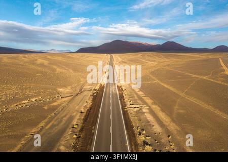 A straight, empty road cuts through the vast, arid landscape of the Atacama Desert in the Chilean Altiplano, offering a breathtaking view of rugged be Stock Photo