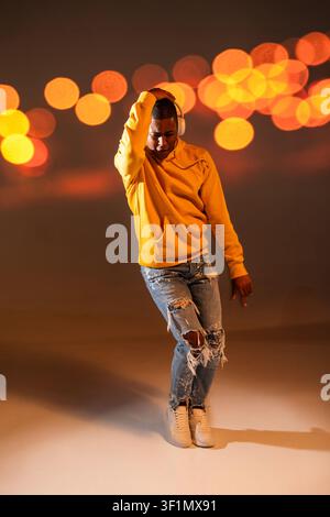 Full-length studio photo of a black man urban dancer wearing a yellow hoodie and ripped jeans, posing with headphones against warm bokeh lights, moder Stock Photo