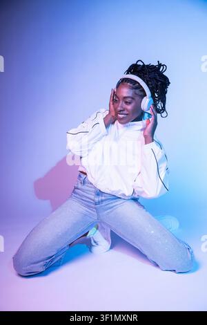 Afro woman dancer kneels in a modern studio, smiling while holding headphones and moving to the beat. Colorful blue and pink lighting creates a vibran Stock Photo
