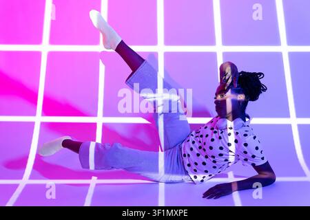 Afro woman dancer poses on studio floor with raised leg, wearing polka dot shirt and pants, lit by vivid pink and purple neon grid projection for a mo Stock Photo