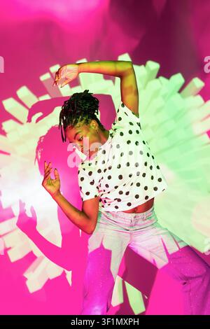 Afro woman dancer striking a dynamic pose under neon pink and green light, creating dramatic shadows on the wall. Modern street style with polka dot s Stock Photo