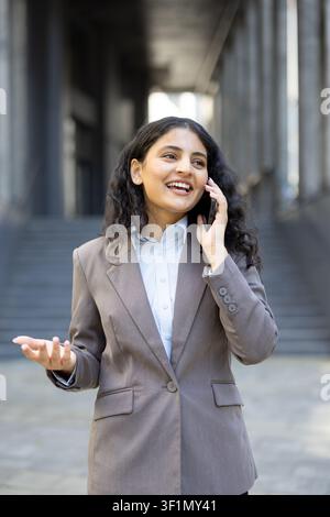 Young professional woman smiling and talking on a smartphone, having an engaging conversation while standing outdoors near a modern building entrance Stock Photo
