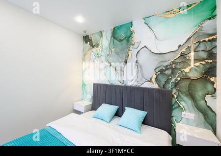 modern bedroom features a bed with a gray headboard, light blue pillows, and a blue throw. The wall behind the bed has a vibrant, abstract, green and Stock Photo