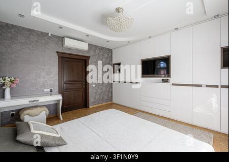 Bright bedroom with white wardrobe & desk, floral wallpaper, wooden door, and a modern bed. Chandeliers hanging from the ceiling Stock Photo