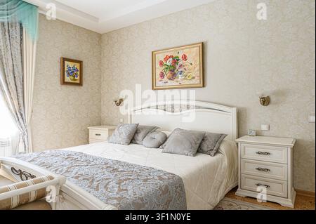 cozy bedroom with a white bed adorned with patterned pillows and a matching bedspread. Artwork hangs above the bed and on the side wall Stock Photo