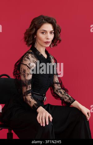 Young woman showcases her chic black lace blouse while posing gracefully in red studio Stock Photo