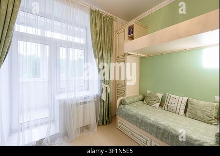 bright bedroom with green walls, sheer white curtains, and patterned green drapes. A bed sits beneath a built-in cabinet, adorned with decorative pill Stock Photo