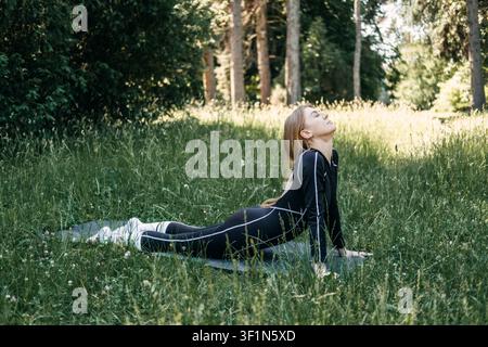 Woman performing cobra pose on yoga mat in grassy field, spine strengthening, morning yoga routine, outdoor exercise Stock Photo
