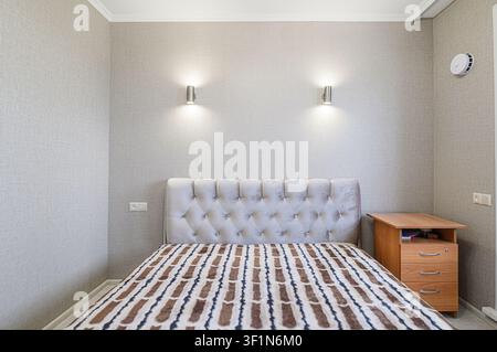 Cozy bedroom scene with a tufted headboard, patterned bedding, bedside table, wall sconces, and neutral wallpaper. Calm and inviting Stock Photo