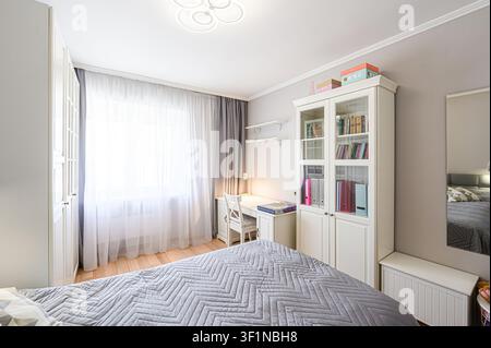 bright, tidy bedroom with light walls, a large window with sheer curtains, white furniture, and a bed with a grey patterned duvet Stock Photo
