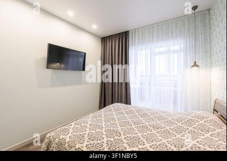 Modern bedroom with a bed, TV on the wall, curtains, and a hanging lamp.  The room is bright and cozy Stock Photo
