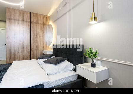 Cozy bedroom with a gray bed, white bedding, wood closet, and modern lighting. A small plant adorns the nightstand Stock Photo