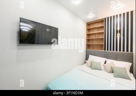 modern bedroom features a TV on a white wall, a grey bed, a bookcase, and black and white striped wall decor Stock Photo