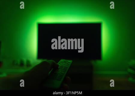 Hand holding a TV remote control pointing at a blank black screen of a television with vibrant green LED ambient backlight in a dark room. Concept of Stock Photo