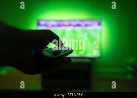Hand silhouette holding a remote control pointing at a blurred TV screen displaying a live soccer match with bright green LED ambient backlight. Great Stock Photo
