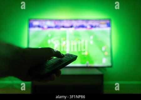 Hand holding a TV remote controller close-up, watching a soccer football match broadcasting on a large screen with vibrant green LED ambient backlight Stock Photo