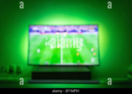 Blurred TV screen showing a live football match with a green grass field and players. Vibrant ambient LED bias backlight on the wall. Great abstract b Stock Photo