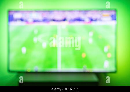 Blurred TV screen showing a live football match with green grass field and players. Vibrant ambient backlight. Great abstract background for sports ba Stock Photo