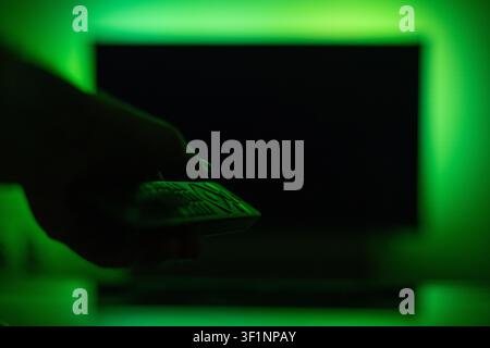 First-person perspective of a hand with remote pointing at a turned-off TV screen illuminated by bright green ambient LED bias lighting. Modern smart Stock Photo