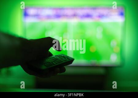 Watching a live soccer tournament at home. Male hand holding a black remote control in front of a big TV screen with a vibrant green LED light backgro Stock Photo