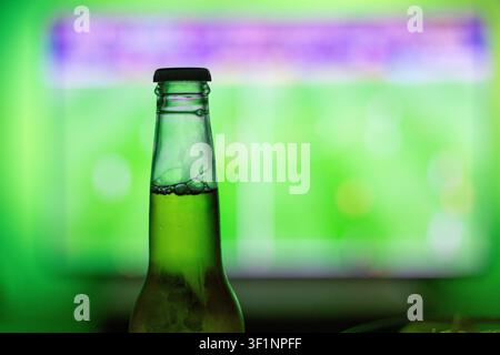 Close-up of a cold beer bottle with a blurred TV screen showing a live football match in the background. Vibrant green ambient backlight. Perfect for Stock Photo
