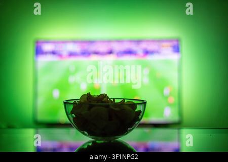 Glass bowl of crispy potato chips on a reflective table with a blurred TV screen showing a football match in the background. Green ambient backlight. Stock Photo
