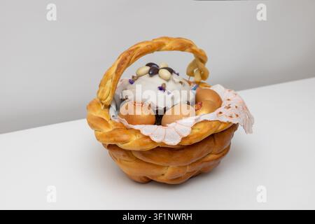 Golden braided bread basket holds decorated eggs sweet bread topped with white icing edible flowers. Soft lighting highlights warm tones against white Stock Photo