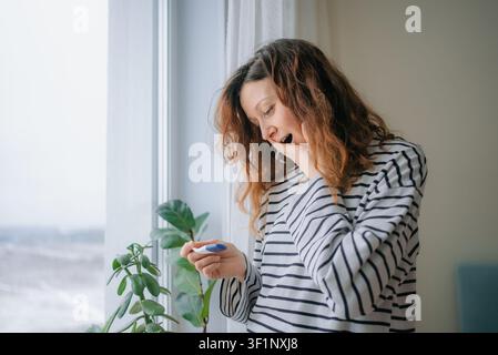 Young woman receiving an unexpected positive pregnancy test result, experiencing shock and disbelief near a window, contemplating her future and motherhood journey Stock Photo