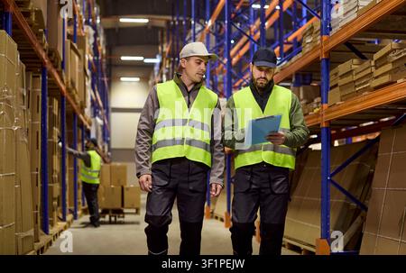 Warehouse workers inspecting inventory on racks, discussing logistics tasks Stock Photo