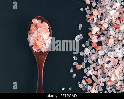Coarse pink himalayan salt crystals in a dark wooden spoon and scattered on black slate stone background. Top view flat lay composition with copy space. Perfect for cooking, spa or wellness concept Stock Photo