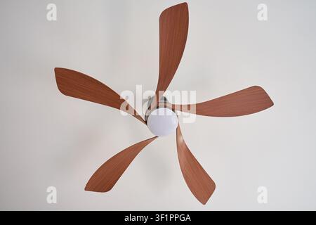 Ceiling fan with five wooden blades and integrated light mounted on white ceiling. Concept of home cooling and air circulation Stock Photo